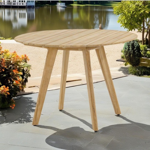 15916 Surf  42 inch Round Teak Dining Table on concrete patio surrounded with shrubs and trees with lake and dock house in background