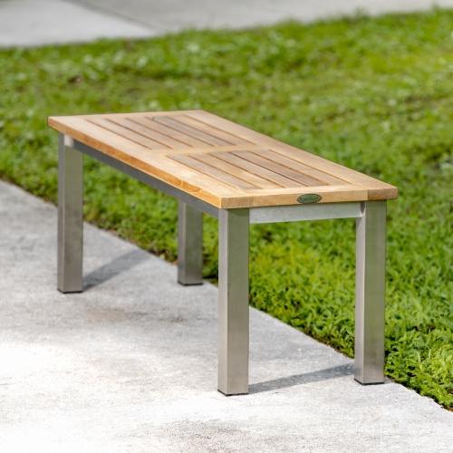 23940 Vogue teak and stainless steel 4 foot backless bench angled end view on concrete walkway with grass in background