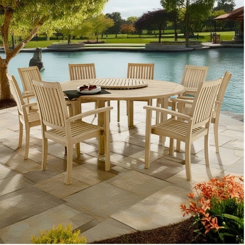 70154 Buckingham 9 piece teak Dining Set of 4 armchairs and round teak dining table angle view on patio with flowering plants and lake and green grass in  background