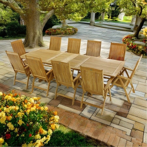70205 Veranda Barbuda 11 pc Teak Dining Set of Grand Veranda Rectangular Table 10 reclining Barbuda chairs on stone paver in a garden courtyard surrounded by trees and flowering shrubs 