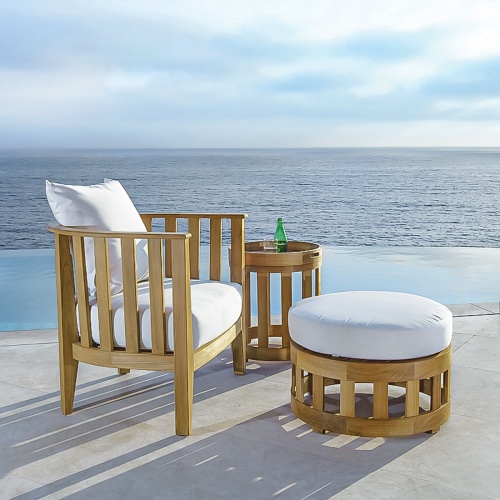 70257 kafelonia teak three piece set showing club chair side table with glass and bottle sparkling water and ottoman with cushions on patio with pool and ocean in background