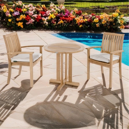 70296 Sussex 3 piece Teak Bistro Set of 2 teak dining armchairs with optional seat cushions and round 30 inch diameter dining table side angled on stone deck next to pool with flowering shrubs in background