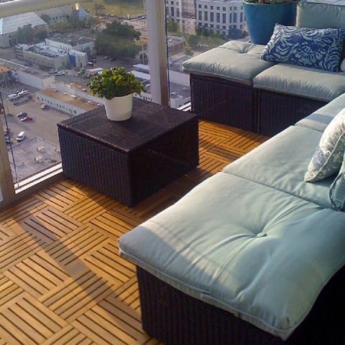 70409 parquet teak tiles one hundred cartons covering nine hundred ten square feet on condo balcony with glass railing potted palm and flowering plant a sofa set overlooking a city view 