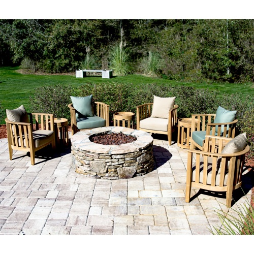70421 kafelonia nine piece teak fire pit conversation set with five cushioned lounge chairs and four teak side tables with tray top on stone patio with  grass shrubs and trees in background 