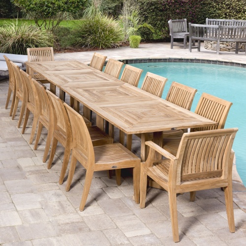 70436 Grand Pyramid 15 piece teak Dining Set of 2 armchairs 12 side chairs a teak rectangular table on paver patio next to pool with landscape plants grass lawn and shrubs in background
