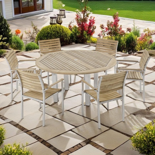 70442 Vogue 7 piece teak and stainless steel Dining Set of 6 armchairs and a round 60 inch diameter table angled on stone patio surrounded by plants with grass lawn in background 