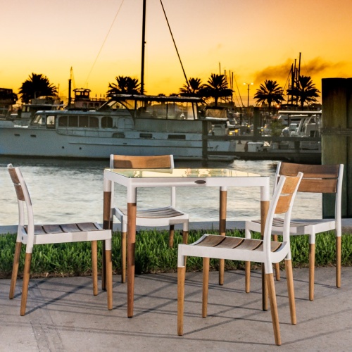 70449 Bloom Glass Top Dining Set of Bloom teak and powder coated aluminum 32 inch square dining table on concrete walkway with boat dock and marina with sunset sky in background