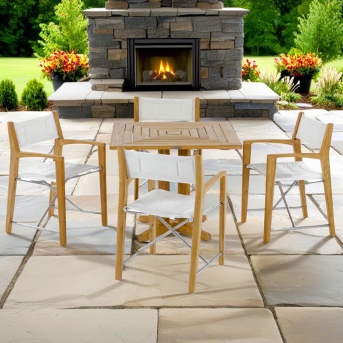 70455 Odyssey Pyramid 5 pc Bistro Set of square teak table and 4 folding teak director chairs on large stone paver patio with fireplace and trees in background
