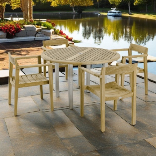 70478 Vogue Horizon Dining Set of Vogue round teak and stainless steel dining table and 4 Horizon teak chairs side angled on stone patio with boat in background