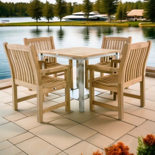 70481 Vogue Veranda teak dining Set of Vogue Square teak and stainless steel dining table and 4 Veranda armchairs side angled view on paver with lake in  background