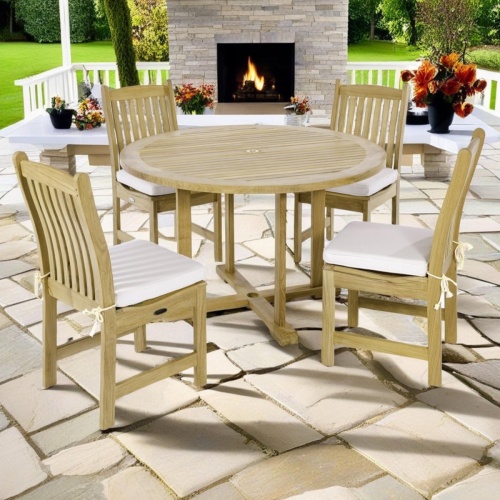 70484 Veranda 4 foot round Dining Set of 4 teak side chairs with optional seat cushions and 4 foot round teak dining table on stone patio with fireplace plants and grass in background