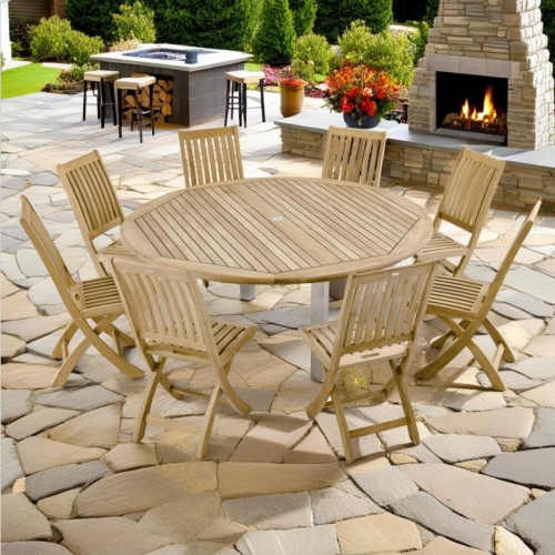 70486 Vogue Barbuda 9 pc Side Chair Set of a Vogue Teak & Stainless Steel  Dining Table and 8 Barbuda Folding Chairs angled on stone patio with fireplace trees and landscaping plants in the background