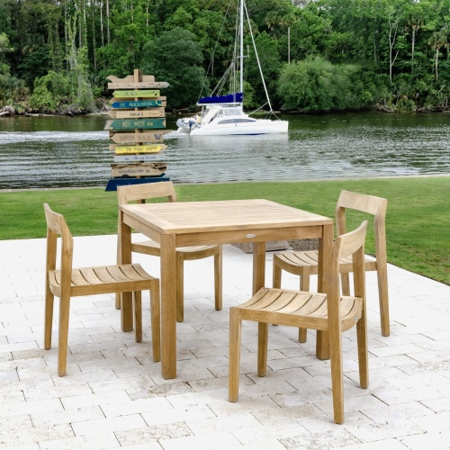 70494 Horizon 5 piece Teak Dining Set of 4 teak side chairs and a 36 inch square teak dining table on concrete patio with grass trees and yacht in background