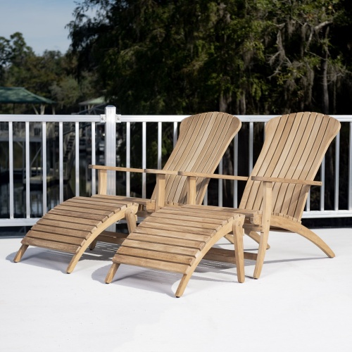 70509 Double Adirondack Set of 2 teak chairs and 2 teak foot rests on concrete patio showing metal balcony rail trees boathouse dock and water in background