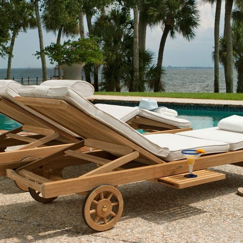70510 Somerset teak double chaise set with two folded towels and a hat on river rock patio facing pool with palm trees ocean view blue sky in background