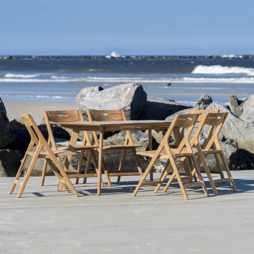 70518 Surf 7 piece teak folding Dining Set on beach with large boulders in behind dining set with ocean and blue sky background
