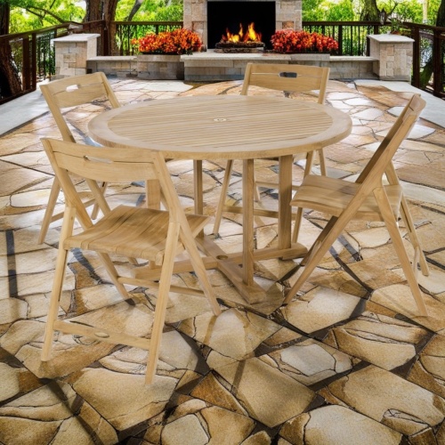 70534 Surf Teak 4 ft Round Dining Set of a teak 48 inch round table and 4 folding chairs side angled on terrace surrounded by plants and trees and fireplace in  background 