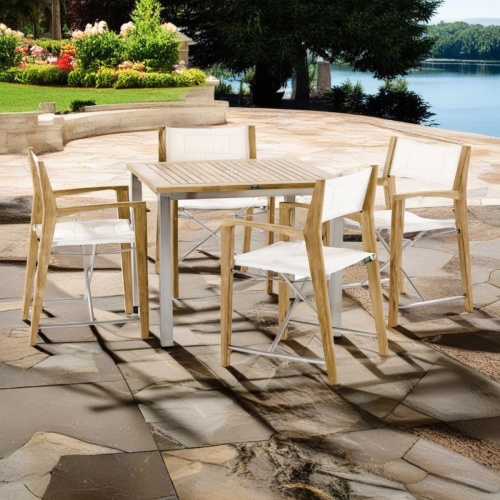 70539 Vogue Odyssey Dining Set of Vogue teak and stainless steel square dining table and 4 Odyssey Chairs angled view on patio with landscape trees & Shrubs with lake in background