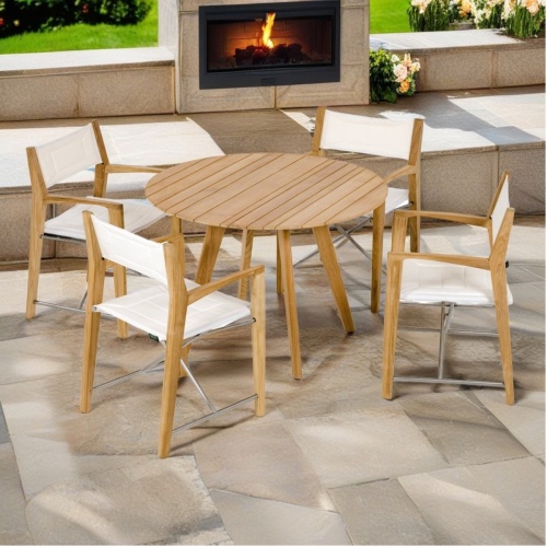 70543 Odyssey Surf Dining Set of a teak 42 inch round dining table and 4 Odyssey Chairs side angled view on stone patio with outdoor fireplace and flowering plants in background