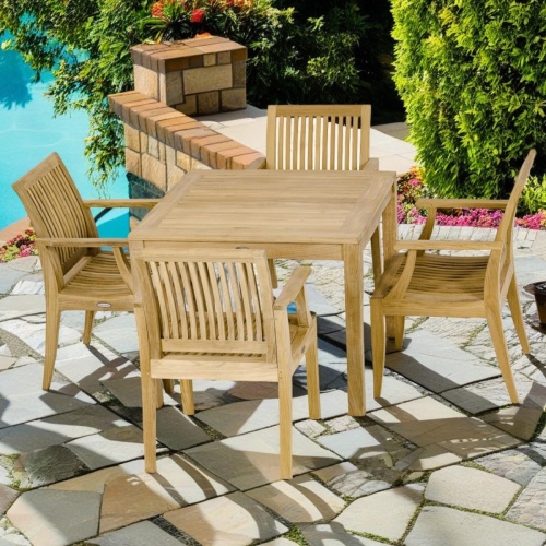 70555 Laguna 5 pc Square Teak Dining Set of a teak 36 inch square dining table and 4 dining chairs on stone patio overlooking a pool with trees and plants in background