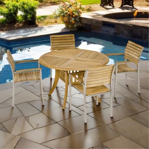 70581 Grand Hyatt Vogue Dining Set of round 48 inch diameter teak dining table and 4 Vogue Side Chairs on pavers in front of pool with landscape shrubs in the background