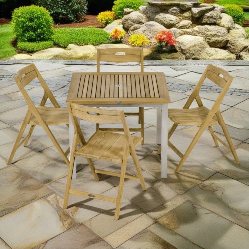 70589 Vogue Surf 5 pc Dining Set of teak table and 4 folding side chairs on stone patio with landscape plants and grass lawn in background 