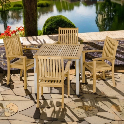 70590 Vogue Sussex Dining Set of Vogue 36 inch square teak and stainless steel dining table and 4 Sussex Chairs angled view on patio with lake and landscape trees in background