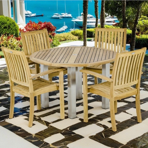 70624 Vogue Sussex Teak Dining Set of a teak and stainless steel 48 inch round dining table and 4 Teak Stacking Chairs side angled view on stone patio overlooking a harbor in background