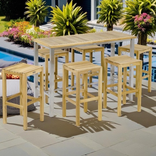  70630 Vogue Somerset 7 piece Backless Bar Set of Vogue Bar Table and 6 teak backless stools on pool deck surrounded by landscaping shrubs with house in background 