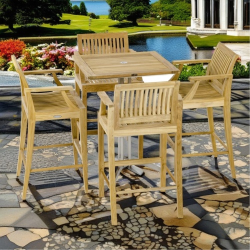 70637 Laguna Vogue Square Bar Set of a teak and stainless steel 30 inch square bar table and 4 teak barstools with armrests side view on stone patio with pool conservatory in background