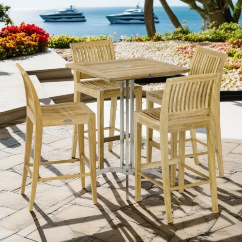 70638 Laguna Vogue 3 piece Teak Square Bar Set of 36 inch square teak and stainless steel bar table and 2 teak side barstools on terrace surrounded by plants and trees overlooking ocean with yachts in background