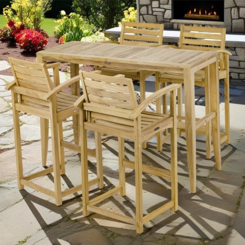 70647 Somerset Laguna Teak Bar Set of teak rectangular Bar Table and 4 Bar Stool with arms angled on pavers surrounded by plants and stone fireplace in background