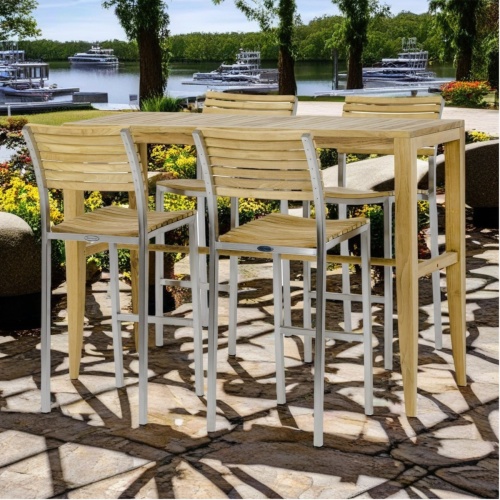 70648 Laguna Vogue Bar Set of Laguna teak rectangular table and 4 Vogue Barstools angled on pavers surrounded flowering plants and trees with boats docked on waterway in background 