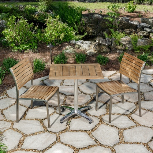 70665 Vogue Bistro Set of a teak and stainless steel rectangular table and 2 teak and stainless steel side chair on stone patio with landscape plants in background