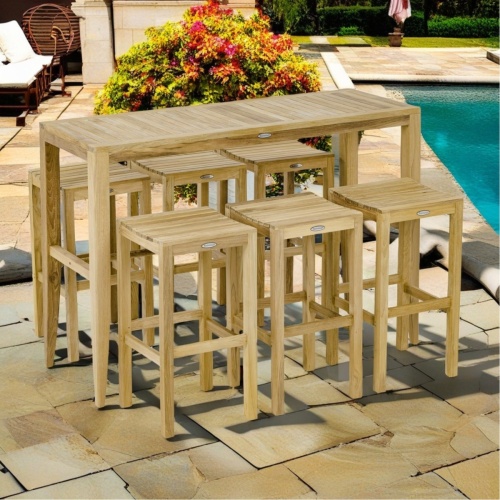 70680 Laguna 5 foot Teak Rectangular Bar Table and 6 Somerset Backless Barstools angled view on concrete patio with pool and landscaping plants and grass lawn in background. 
