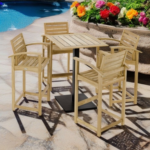 70693 Somerset 5 piece High Bar Patio Set of a 30 inch square teak table in bar height and 4 teak barstools angled on a stone pool deck next to a concrete planter with flowers