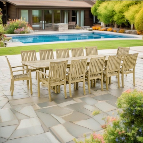 70757 Sussex Veranda teak 13 piece dining set of 12 armchairs and rectangular extension dining table angled on pavers with landscape plants and pool and house in background
