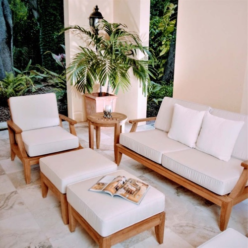 70877 Laguna 7 piece teak Love Seat Set on travertine tile patio open magazine on ottoman pillar candle in holder on side table potted palm tree in corner porch light with tree background