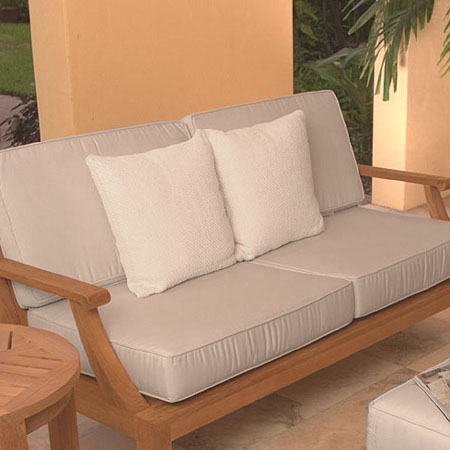 73122LM Laguna teak Sofa Cushions on Laguna Sofa next to round side table on tile patio against wall with grass lawn and trees in background