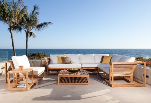 11800RFDP Maya teak lounge set with bowl of apples a potted plant and magazine on table shelf on concrete patio surrounded by two palm trees and plants with ocean and blue sky background