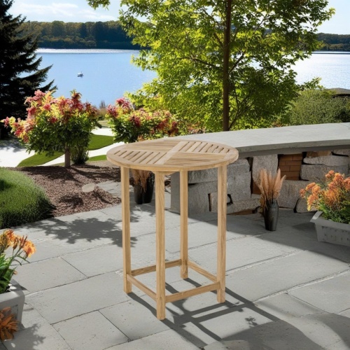 15334 Somerset Teak Round 36 inch diameter Bar Table angled view on stone patio with landscaping plants and lake view in background