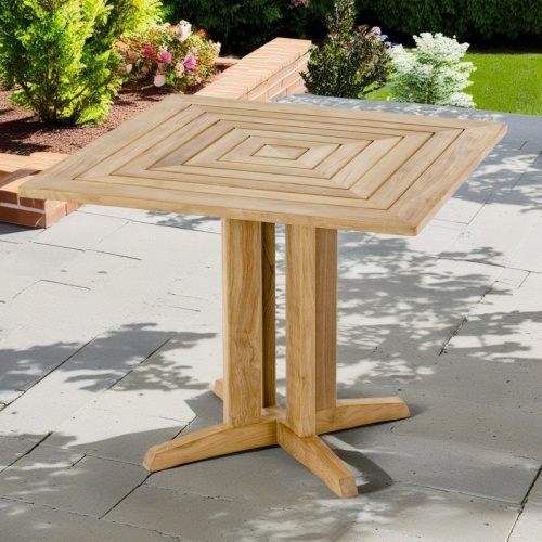 15815 Square 36 inch Pyramid Teak Table angled view on concrete patio surrounded by landscaping plants and grass lawn in background  