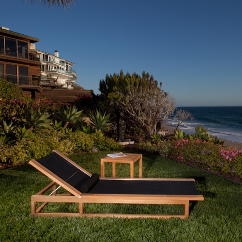 16771 Maya Chaise Lounger in black Textilene mesh fabric and side table with magazine on a green lawn overlooking the ocean with two homes and flowering plants and trees in background 