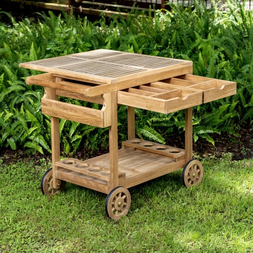 17105 Alicante teak Trolley Cart on grass showing fold out top 2 pull out drawers and 8 wine bottle holder on bottom shelf and rubber lined wheels with shrubs in background