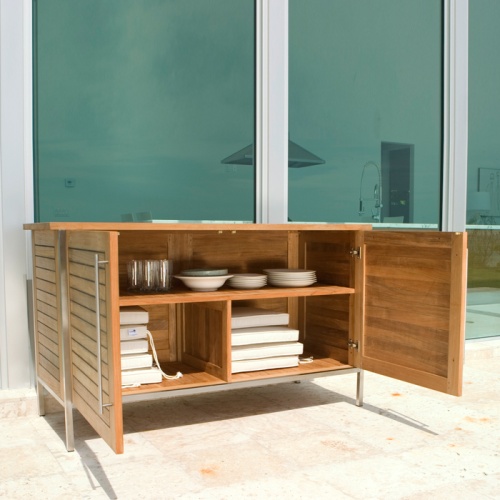 28225RF Vogue teak and stainless steel Sideboard on outdoor patio angled view with sideboard doors open displaying storage area with dishes and seat cushions against glass windows