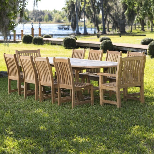 70008 Montserrat Veranda 11 piece oval dining set angled side view on a grass field with palm trees shrubs a boardwalk with 2 lamp posts boat dock and lake in background