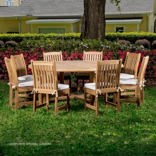 70020 Buckingham Veranda 9 piece teak round dining set with optional seat cushions on grass with flowering shrubs trees and landscaping plants with yellow house in background