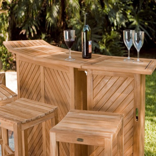 70084 somerset teak five piece bar set showing three wine glasses and wine bottle on top with stone patio a grass area paver walkway and lush tropical plants in background
