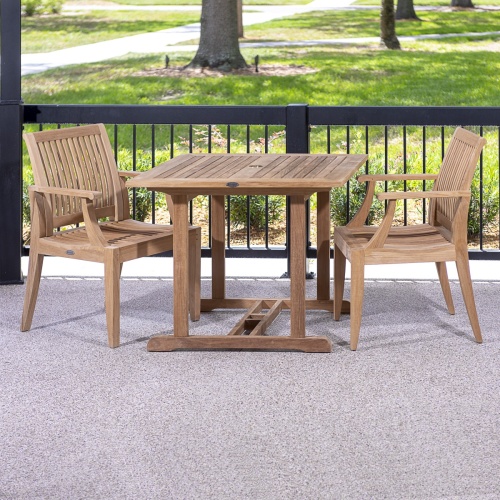 70301 Laguna 3 piece teak Dining Set of a 36 inch square dining table and 2 teak dining armchair on concrete patio against a black railing with grass lawn and trees in background