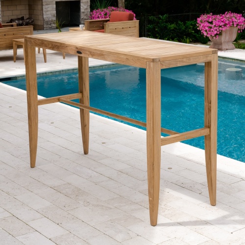 70680 Laguna 5 foot Teak Rectangular Bar Table angled view on concrete patio with pool and fireplace in background